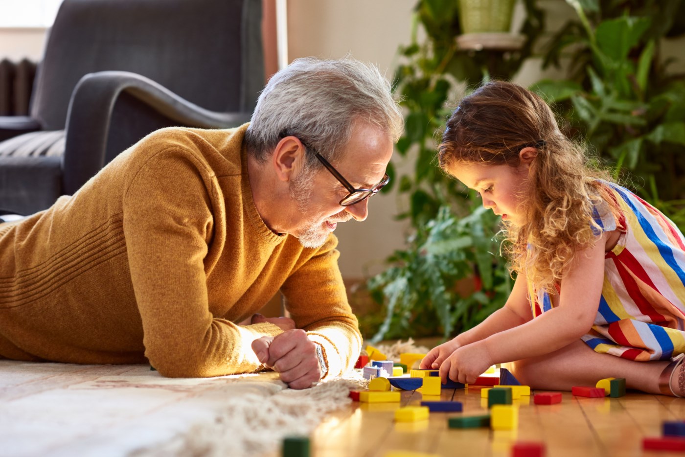 En eldre mann med grått hår og briller bygger Lego på gulvet sammen med sitt barnebarn med krøllete hår i en stripete, fargerik kjole.